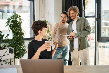 young man with coffee to go pointing at laptop and looking at coworkers while discussing project
