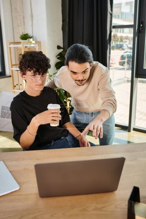 joven apuntando a la computadora portátil mientras explica su idea de proyecto a su compañero de trabajo con café para ir