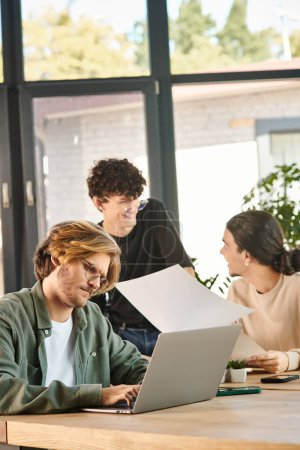 Photo for Young man using laptop, team of young entrepreneurs engaged in project planning in office - Royalty Free Image