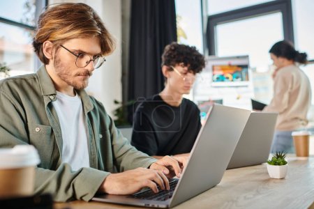 Foto de Equipo joven de startup profundamente enfocado mientras trabaja en computadoras portátiles en el espacio de coworking moderno, hombres de 20 años - Imagen libre de derechos
