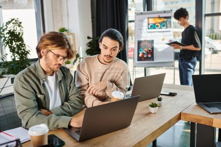 Equipo joven de startup discutiendo proyecto mientras trabaja en computadoras portátiles en un espacio de coworking, hombres de 20 años