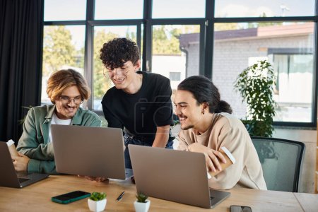 Téléchargez les photos : Souriants jeunes hommes dans la vingtaine analysant le projet ensemble sur des ordinateurs portables dans un espace de coworking ensoleillé, démarrage - en image libre de droit