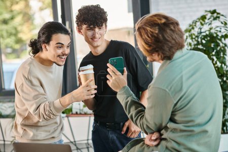 young coworkers sharing laugh during coffee break in a bright office, man in 20s showing smartphone