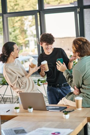young coworkers smiling during coffee break in a bright office, man in 20s showing smartphone