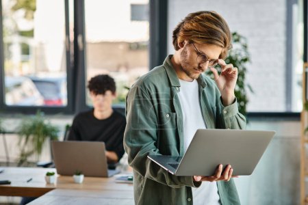 Téléchargez les photos : Jeune homme d'affaires confiant dans des lunettes utilisant un ordinateur portable et debout dans un espace de coworking moderne - en image libre de droit