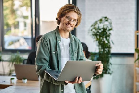 alegre joven hombre de negocios en gafas usando el ordenador portátil y de pie en un espacio de coworking moderno