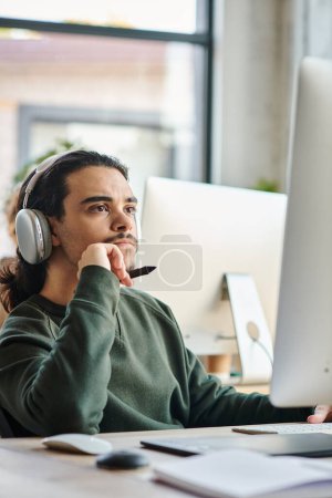 Téléchargez les photos : Professionnel masculin réfléchi dans les écouteurs tenant stylet stylo et travaillant sur le projet à son bureau - en image libre de droit