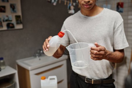 cropped african american man pouring chemical solution in measuring cup, analog film development