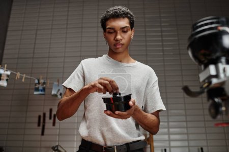 Foto de Joven fotógrafo afroamericano asegurando tapa de lata de película en cuarto oscuro, desarrollo analógico - Imagen libre de derechos