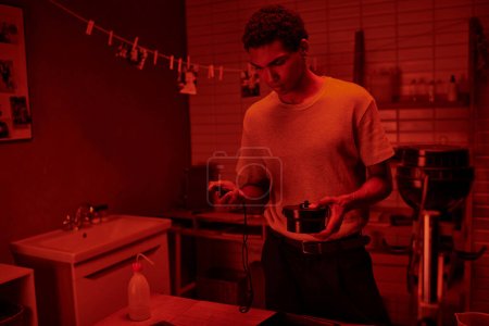Fotograf in rot beleuchtetem Raum, schwarzer Mann steuert Filmentwicklung vorsichtig mit Dunkelkammer-Timer