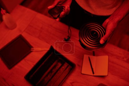 zugeschnittener Fotograf in rot beleuchteter Dunkelkammer, schwarzer Mann mit Timer kümmert sich sorgfältig um Filmentwicklung