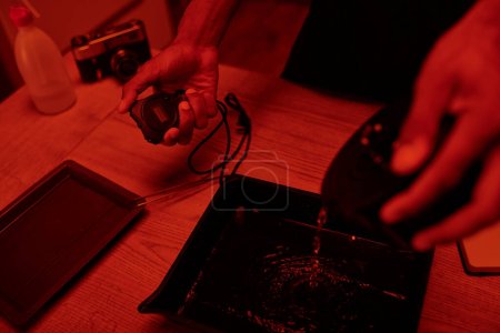 Fokus auf Timer in der Hand des Fotografen gießt Entwickler in Tablett zur Filmbearbeitung, rote Dunkelkammer