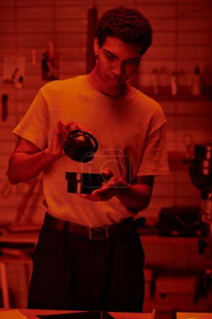 Foto de Fotógrafo guapo sosteniendo bote de película y desarrollando fotos en cuarto oscuro con luz roja - Imagen libre de derechos