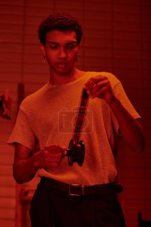Foto de Fotógrafo afroamericano enfocado desenrolla película en preparación para el desarrollo en el cuarto oscuro rojo - Imagen libre de derechos