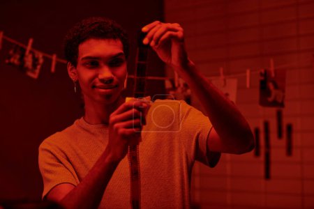 Foto de Feliz afroamericano fotógrafo examina desarrollado tira de película, bañado en cuarto oscuro con luz roja - Imagen libre de derechos