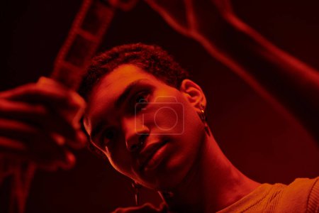 foco en el fotógrafo afroamericano examina tira de película desarrollada, bañado en el cuarto oscuro rojo
