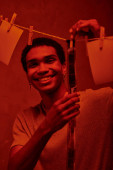happy african american man hanging freshly developed film strip  in a red-lit darkroom, nostalgia t-shirt #692601298