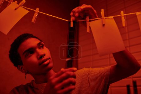 photographe afro-américain regardant développé bande de film dans une chambre noire éclairée en rouge, nostalgie