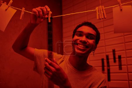 cheerful african american man hanging freshly developed film strip  in a red-lit darkroom, nostalgia