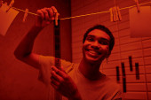 cheerful african american man hanging freshly developed film strip  in a red-lit darkroom, nostalgia t-shirt #692601326