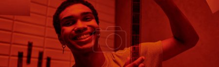 Foto de Alegre afroamericano hombre colgando recién desarrollado tira de película en un cuarto oscuro de luz roja, pancarta - Imagen libre de derechos