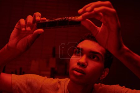 fokussierter afrikanisch-amerikanischer Fotograf beim Betrachten eines entwickelten Filmstreifens in einer rotbeleuchteten Dunkelkammer