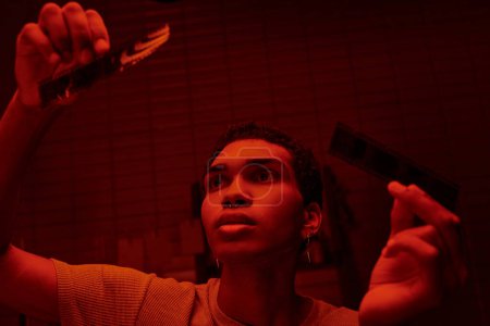 Foto de Fotógrafo afroamericano enfocado comparando tiras de película desarrolladas en un cuarto oscuro iluminado por el rojo - Imagen libre de derechos