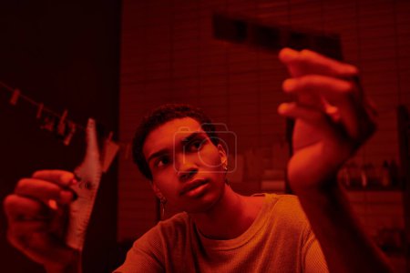 konzentrierter afrikanisch-amerikanischer Fotograf beim Vergleich entwickelter Filmstreifen in einer rotbeleuchteten Dunkelkammer