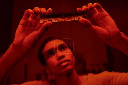 fotógrafo afroamericano enfocado examinar desarrollado tira de película en un cuarto oscuro con luz roja