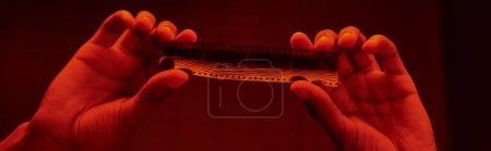 Photo for Cropped banner of african american photographer examine developed film strip in a red-lit darkroom - Royalty Free Image