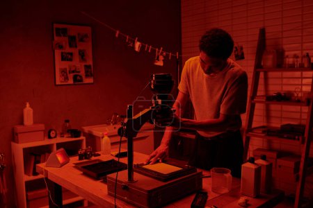 In a red-hued darkroom, a photographer focuses intently on the delicate process of enlarging film