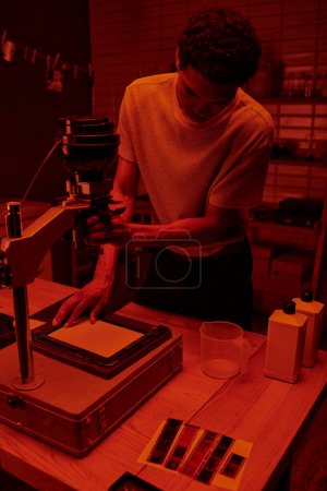 Photo for Dark skinned photographer focuses on delicate process of enlarging film in darkroom with red light - Royalty Free Image