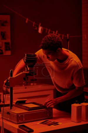 photographe afro-américain inspectant la photo négative sous la lumière rouge de sécurité d'une chambre noire