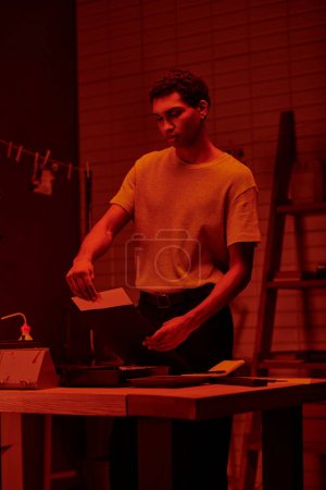 In the glow of red darkroom lights, black photographer prepares photographic paper for printing