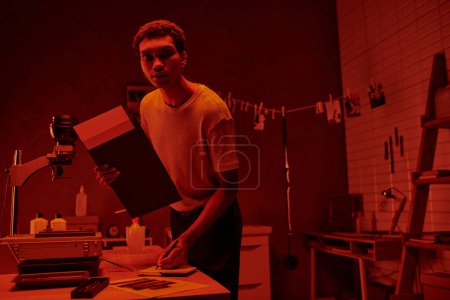 young photographer prepares photographic paper for printing and taking notes in red darkroom