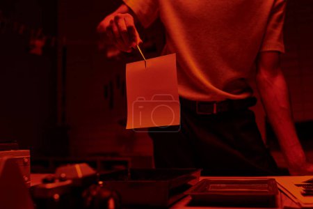 Schnappschuss eines Fotografen mit Pinzette und Fotopapier in einer Dunkelkammer mit rotem Licht