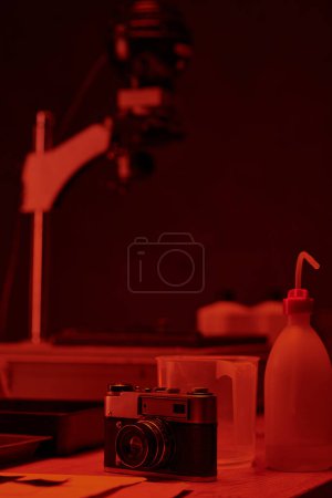 Foto de Cámara analógica y diferentes herramientas para el desarrollo de películas en la mesa en cuarto oscuro con luz roja - Imagen libre de derechos