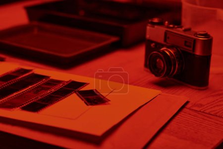 Téléchargez les photos : Une table avec caméra analogique et bandes de film sous l'éclat de la lumière rouge dans la chambre noire, intemporelle - en image libre de droit