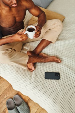 Photo for African american man in pajama pants sitting on bed with coffee cup near smartphone, cropped shot - Royalty Free Image