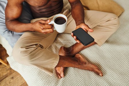 Foto de Hombre afroamericano en pantalones de pijama sentado en la cama con café y teléfono inteligente con pantalla en blanco - Imagen libre de derechos