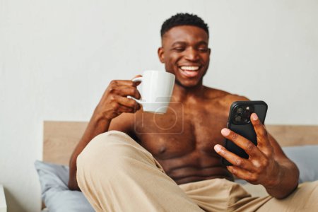 Photo for Joyful athletic african american man with coffee cup browsing internet on smartphone in bedroom - Royalty Free Image