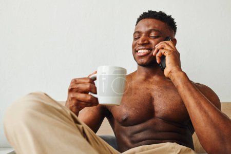 Photo for Cheerful muscular african american man with morning coffee talking on smartphone in bedroom - Royalty Free Image