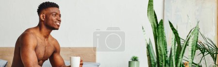 smiling african american shirtless man sitting with coffee cup near green plants in bedroom, banner