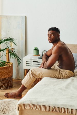 Photo for Thoughtful african american shirtless man in pajama pants sitting and looking at camera in bedroom - Royalty Free Image