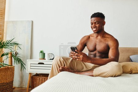 Photo for Joyous and muscular african american in pajama pants browsing social media om smartphone in bedroom - Royalty Free Image