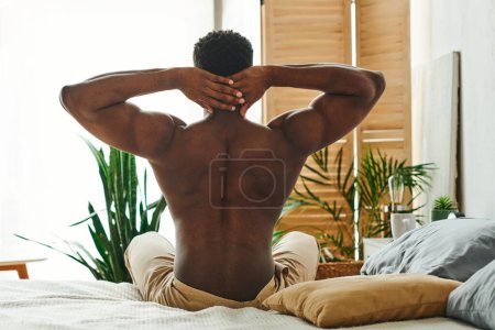 Photo for Shirtless and muscular african american man sitting and stretching on bed in morning, back view - Royalty Free Image
