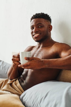 Photo for Smiling african american man with shirtless torso holding coffee cup and looking at camera on bed - Royalty Free Image