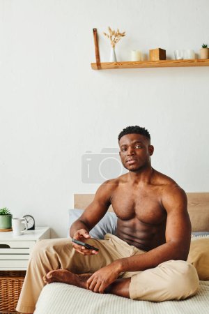 Photo for Muscular african american man in pajama pants sitting on bed with smartphone and looking at camera - Royalty Free Image