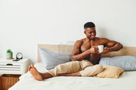 Photo for Joyful shirtless african american man in pajama pants sitting with coffee cup on bed in morning - Royalty Free Image