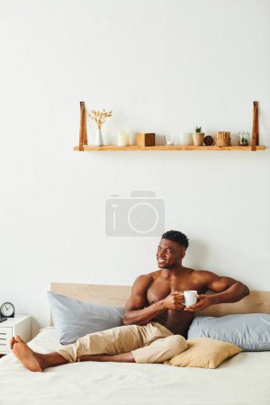 Photo for Cheerful athletic african american man in pajama pants sitting with coffee cup on bed in morning - Royalty Free Image